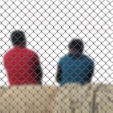 Two men sitting with their backs turned behind a chain-link fence