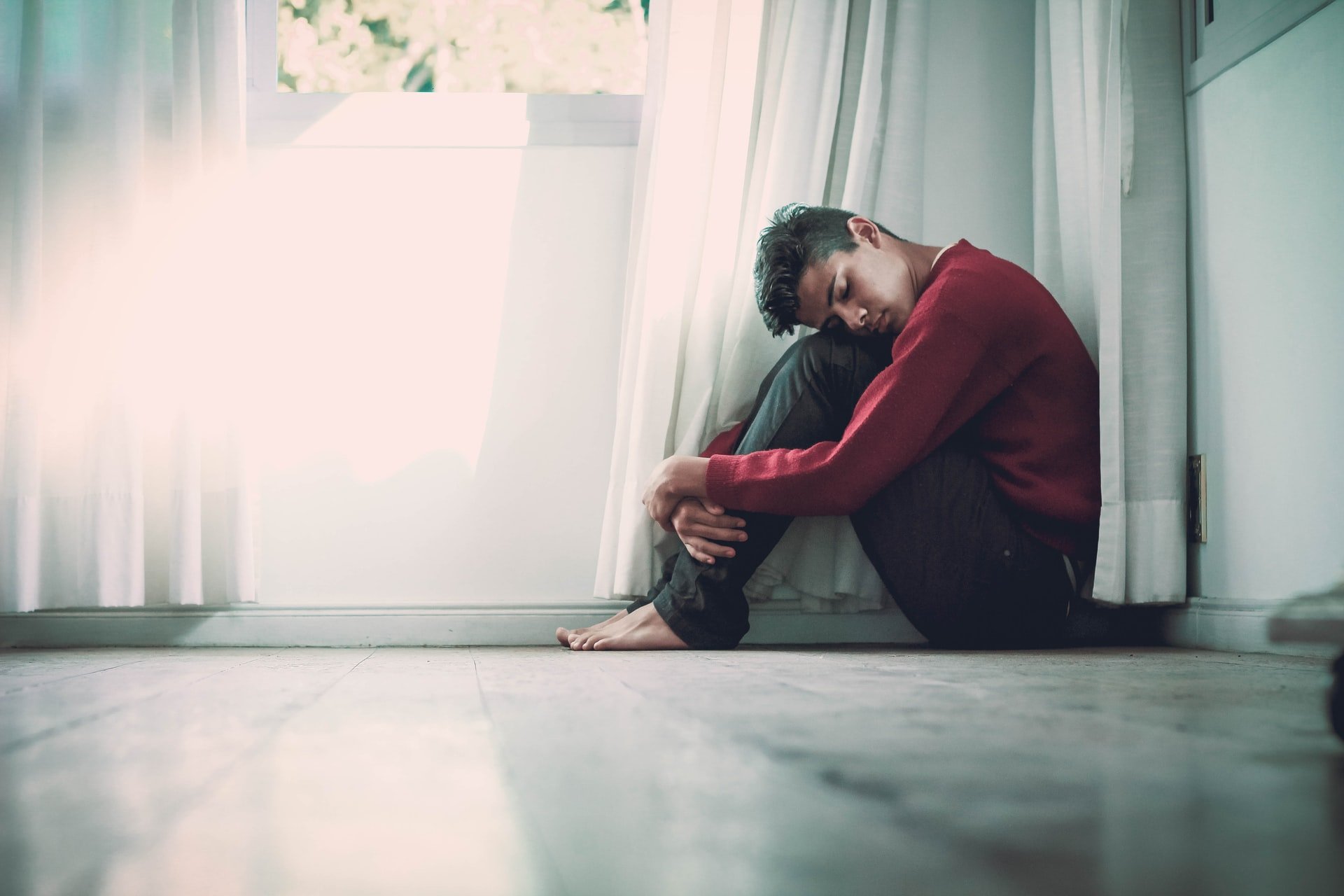 A man looks sad while sitting on the floor next to a window