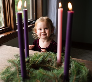 A toddler girl smiles happily at a full lit Advent wreath that sits above her on a table top