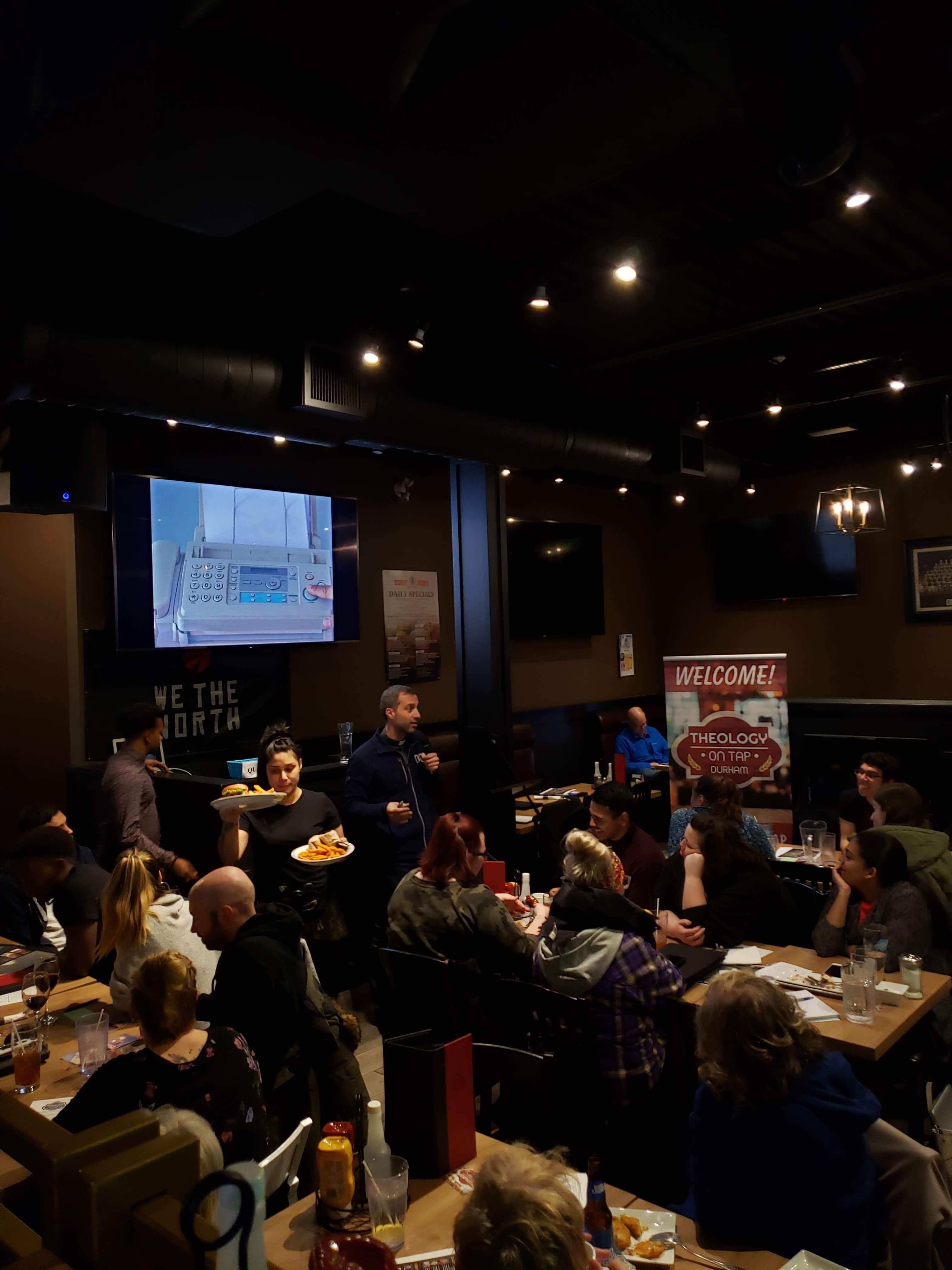 Group of young adults in a pub/restaurant eating and drinking while listening to a guest speaker