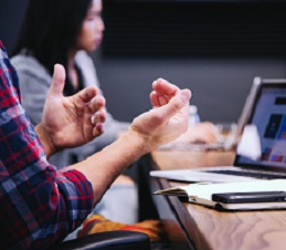 A man's hands in front of a laptop, seemingly in mid-sentence. In a classroom. A woman is seated, looking at a laptop in the background