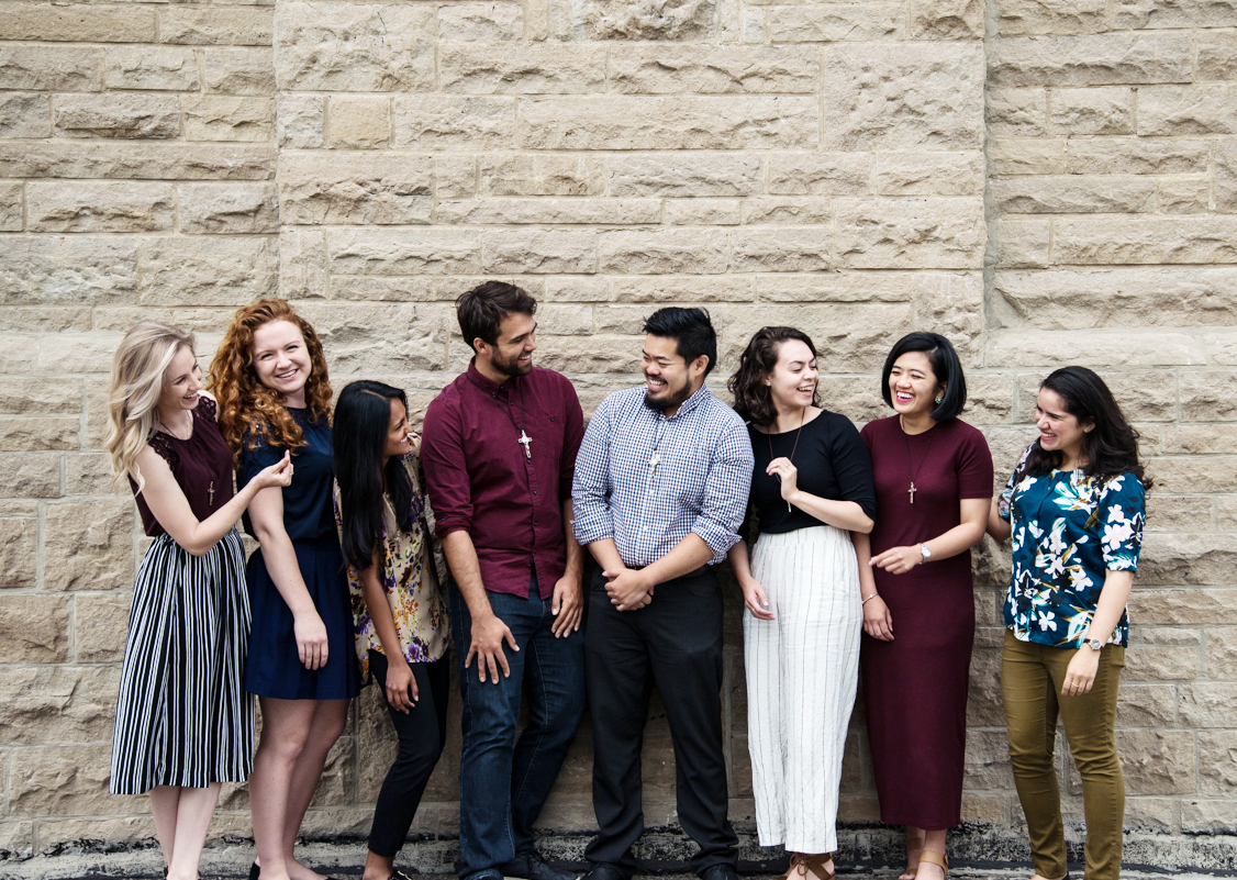 Eight young adults lined up next to a stone wall smiling and laughing at each other
