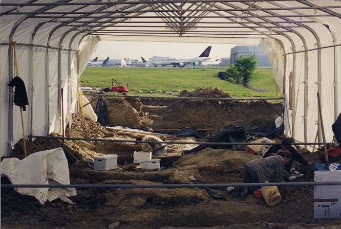 People work at Elmbank Cemetery archaeological site under a canopy. A plane is in the background.
